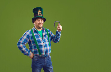 Smiling overweight man in checkered shirt and traditional hat standing and holding horseshoe in hand celebrating Saint Partick day holiday over green wall background. Irish traditional holiday concept