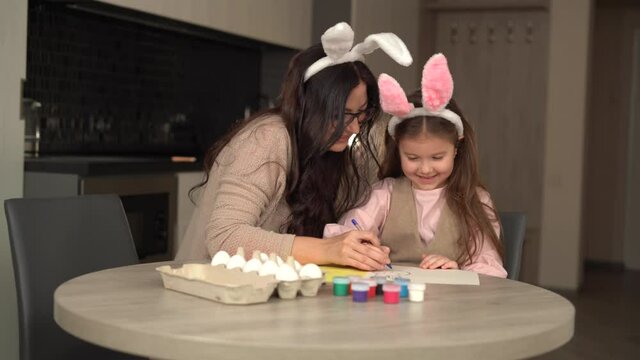 Cheerful family is preparing for Easter holiday mom and daughter are sitting at the table draw a hare on paper.