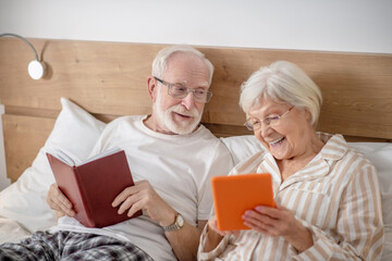 Aged married couple lying in bed and having good time while reading