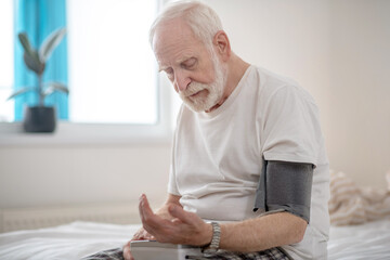 Grey-haired elderly man sitting on a bed and checking his blood pressure