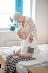 Grey-haired elderly woman helping her husband to check blood pressure