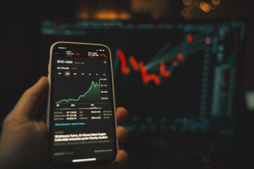 Hand holding a modern smartphone with the stocks market app and big screen blurred on the background