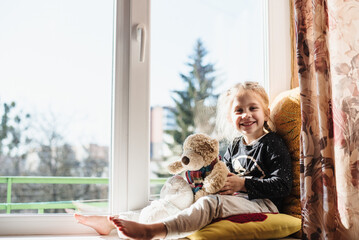Cute little girl enjoying the sunshine while sitting at the window. The child is playing sitting on...