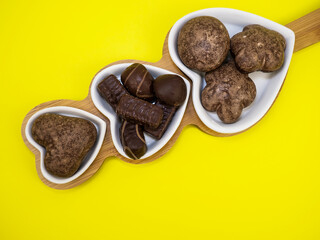 Chocolate candies and gingerbread cookies lie on a wooden board in the form of a heart on a yellow background. Sweets.