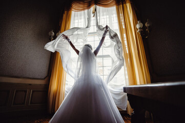 The bride touches the curtains while standing near the palace window. beautiful bride in white dresses opening curtains. Rear view.