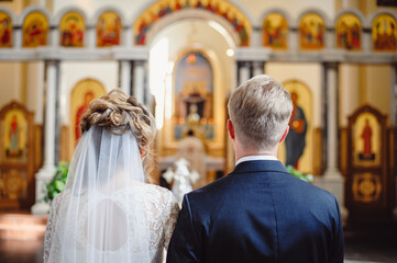 Newlyweds at the wedding ceremony in the church are facing the altar. The priest blesses the bride and groom for a long life. Rear view