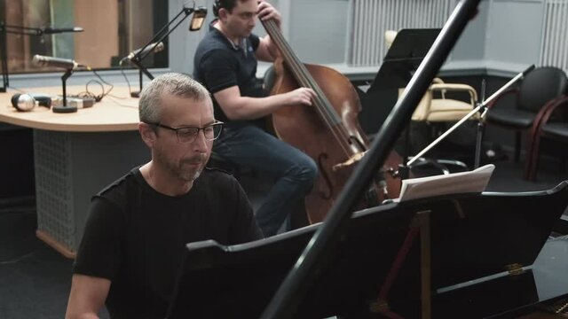 Medium handheld footage of concentrated male caucasian jazz pianist and contrabass player performing instrumental music at professional recording studio full of musical equipment