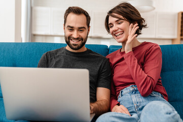 Happy young couple waving hand and using laptop while sitting on couch