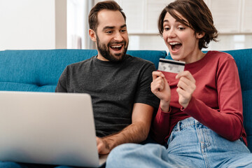 Excited young couple using laptop and credit card while sitting on couch