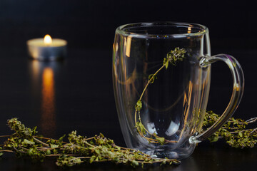 Herbal tea, transparent cup,  burning candle   on black background. Thyme leaves in transparent,   empty glass cup.