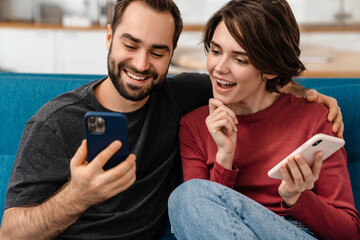 Obraz premium Excited young couple using mobile phones while sitting on couch