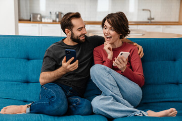 Excited young couple using mobile phones while sitting on couch