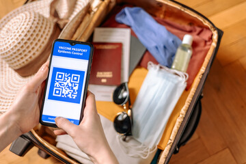 Top view of a female's hands hold a smartphone with an immune digital health passport. In the background, a suitcase with protective masks and gloves, straw hat and sunglasses in a blur.New normal
