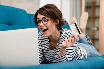 Excited beautiful woman using credit card and laptop on couch