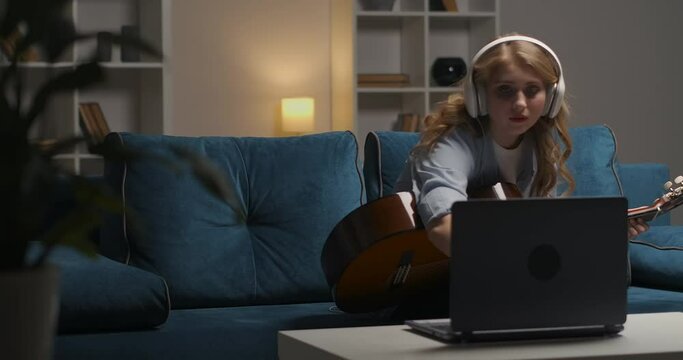 teen girl is playing guitar, viewing notes and tutorial in internet on notebook at evening, spending free time and learninf online