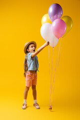 full length of shocked kid in straw hat reaching balloons on yellow
