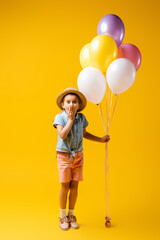 Fototapeta na wymiar full length of shocked kid in straw hat covering mouth and holding balloons on yellow