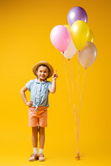 full length of happy kid in straw hat holding balloons and standing with hand on hip on yellow