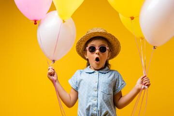 shocked kid in straw hat and sunglasses holding balloons isolated on yellow