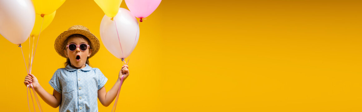 shocked kid in straw hat and sunglasses holding balloons isolated on yellow, banner