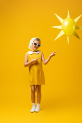 full length of happy girl in headscarf and sunglasses pointing with fingers at decorative sun with balloon on yellow