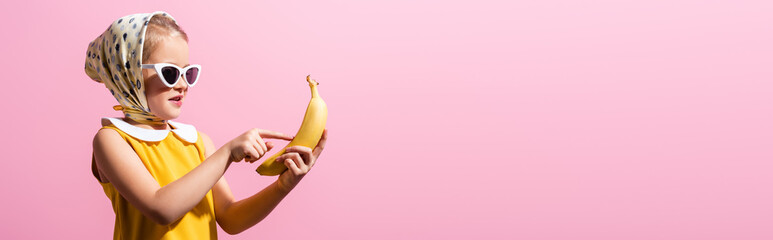 happy girl in headscarf and sunglasses pointing with finger at banana isolated on pink, banner