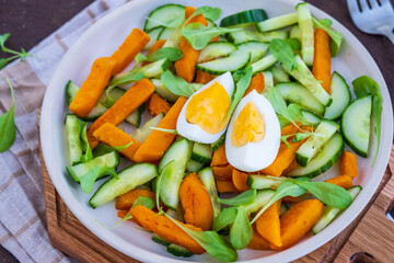 Roasted sweet potato or pumpkin salad, arugula, fresh cucumber and boiled egg on a ceramic plate on a brown concrete background. Sweet potato recipes.