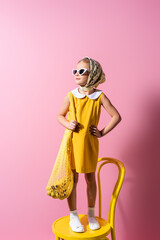 cheerful girl in headscarf and sunglasses holding reusable string bag with bananas while standing on yellow chair on pink