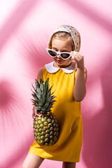 stylish girl in headscarf adjusting sunglasses while holding pineapple on pink