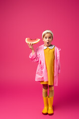 full length of shocked girl in raincoat and rain boots standing with watermelon on crimson