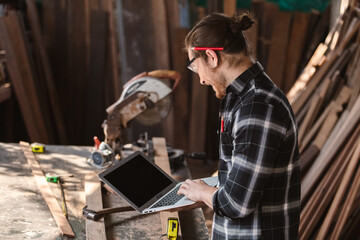 Young carpenter caucasian man worker holding computer laptop at workspace. craftsman profession in wood factory.