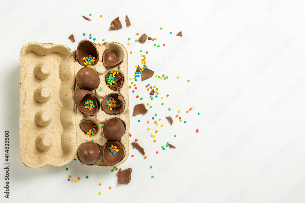 Wall mural Broken Easter Chocolate eggs in a tray and multi-colored decorations on a light background. Easter concept, easter treats,. Banner. Flat lay, top view