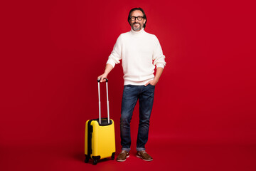 Full size photo of satisfied person hold suitcase hand in pocket toothy smile isolated on red color background