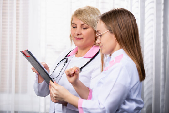 Profile photo of two medical workers discussing assignment sheet. Blond senior doctor, medical chief in white robe showing the assignment sheet to young medical assistant
