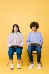 Teen friends using digital tablets on chairs on yellow background