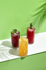 Black currant, orange and raspberry lemonade drink over green background. White table with sunlight and palm leaf hard shadow. Summer, tropical and juicy concept