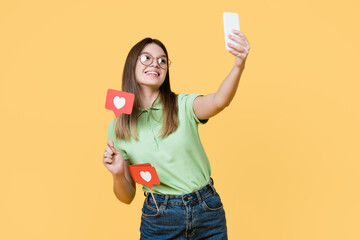 Cheerful teenager holding paper heart on stick and taking selfie isolated on yellow