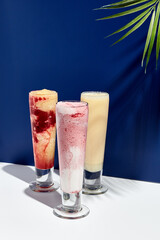 Raspberry and banana, pineapple, strawberry and coconut milk smoothie over white and blue background. Palm leaf with sunshine and hard shadow. Summer, healthy, fresh drink concept