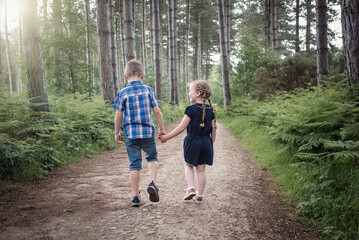 Young boy and girl in shorts holding hands walking through forest woodland on bright sunny summer day in shorts looking at each other love and family lifestyle