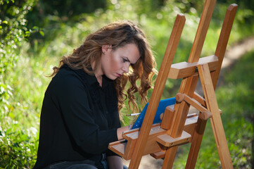 Side view of a portrait of a young white woman drawing a picture on a wooden easel in nature.