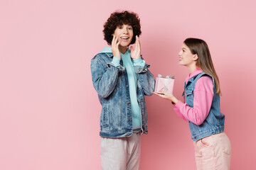 Happy teenager looking at camera near girlfriend with gift isolated on pink