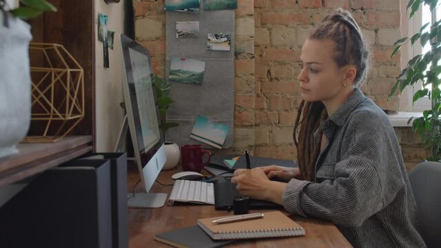 Medium side-view shot of creative female graphic designer with dreadlocks taking picture of computer monitor with sketches drawn using drawing tablet and stylus posting it to social networks