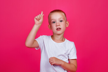 A young little Caucasian guy with blue eyes, wearing a white t-shirt on a pink background, pointing his finger up with a successful idea. Excited and happy.