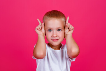 A portrait of a shocked blond boy in white casual clothes stands on a pink background, looking at the camera and pointing up with his index fingers. The surprised boy points to the copy space.