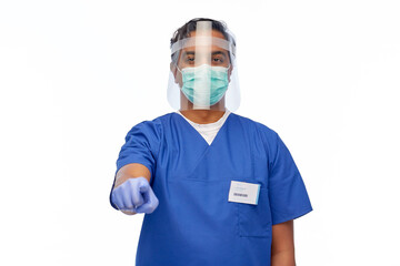 Fototapeta na wymiar medicine, healthcare and protection concept - indian doctor or male nurse in blue uniform, protective medical mask and face shield pointing to camera over white background