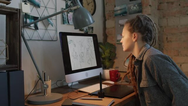 Medium slow-motion shot of young woman with dreadlocks starting drawing sketches via graphic tablet and pen, sitting at table with huge monitor at home