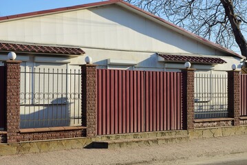 red metal gates and a fence wall of black iron rods and brown bricks in front of the white wall of the house