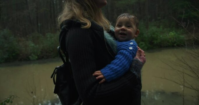 A young mother walking in the woods by water is carrying her baby in a sling