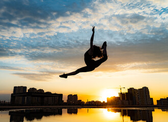 Female flexible dancer jumping during sunset on cityscape background with reflection in the water. Concept of freedom and happiness 