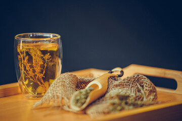 Fototapeta premium Herbal tea flowers in transparent glass cup. Thyme tea's cup and thyme flower on wooden tray. Black background.